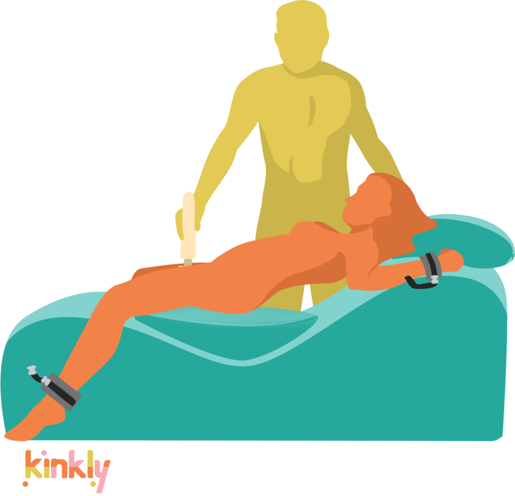 At Your Mercy sex chaise sex position. The receiving partner is laying down on top of the sex chaise, laying almost flat with the chaise cushion underneath the lowest point of the sex chaise. The receiver's wrists and ankles are bound to the sex chaise beneath them and the receiver has a pillow underneath their head. The giving partner is standing next to the sex chaise, holding a wand vibrator between the receiver's thighs. | Kinkly