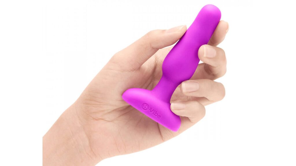 A hand holds the b-Vibe Remote Novice Plug. The plug looks slender and short within the hand. It's slightly thicker than one finger, and it isn't as long as the person's outstretched hand. | Kinkly Shop