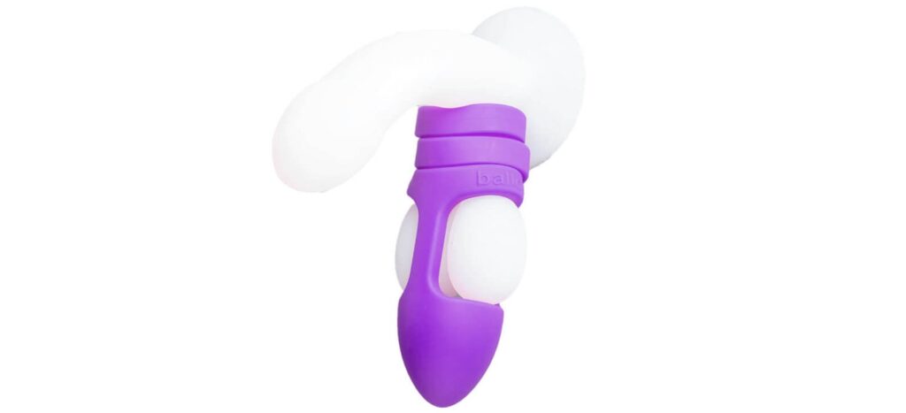 The Balldo in Purple is shown being used on the testicles of a clear dildo. The image shows how the Balldo works - which is by wrapping around the testicles while simultaneously leaving space for the testicles to receive pleasure during penetration. | Kinkly Shop