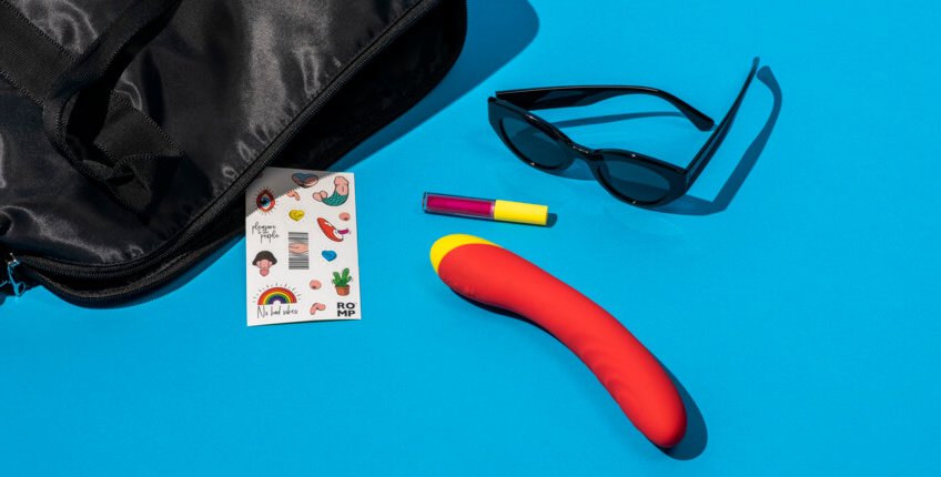 The ROMP Hype G-Spot Vibrator sitting out next to an open bag. The ROMP Hype G-Spot Vibrator is surrounded by a pair of sunglasses, a lip gloss, and a set of ROMP stickers. The vibrator is much thicker than the lip gloss tube and is longer in length than the sunglasses. | Kinkly Shop