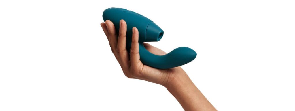 A hand holds the Womanizer Duo 2 at 3/4ths angle. This showcases the air suction hole as well as the upwards curve of the g-spot shaft. The vibrator is longer than it seems, too, with the toy extending beyond the base of the palm. | Kinkly Shop