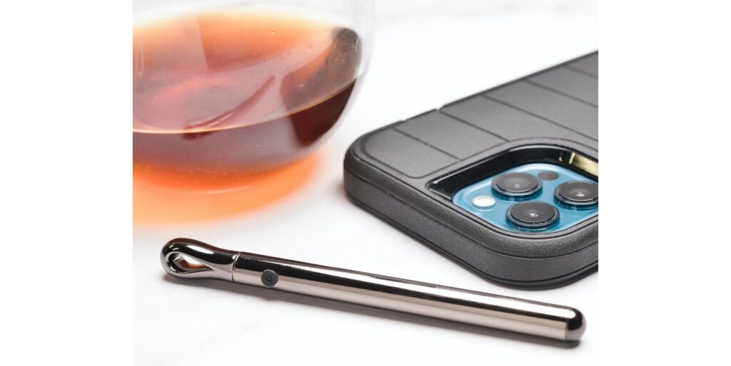 The Evolved Pen Pal sits out in front of a glass of brown liquid and a cell phone. It looks like a small pen laying out on a table. | Kinkly